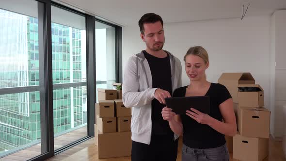 A Moving Couple Looks at the Plan of Their New Apartment on a Tablet - Piles of Cardboard Boxes