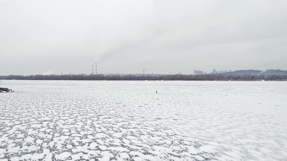 Flying Over a Frozen River with Smoking Chimneys on the Bank and People on the Ice