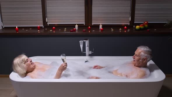 Senior Couple in Love Lying in Warm Bath with Bubbles, Enjoying Relaxation, Drinking Champagne