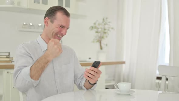 Middle Aged Man Celebrating Success on Smartphone at Home 