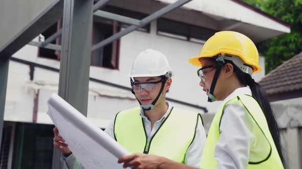 Asian male and female builder engineers wear helmet, holding drawing working on construction side.