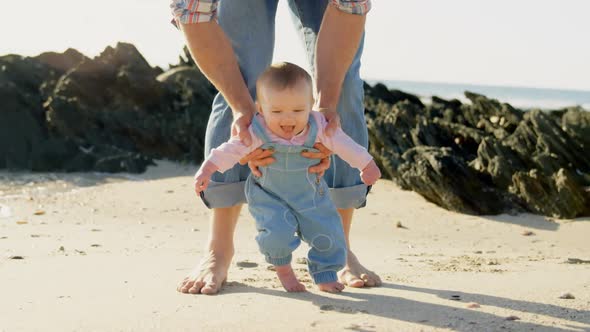 Front view of mid adult caucasian father helping baby to walk at beach on a sunny day 4k