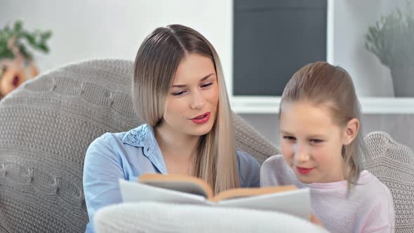 Beautiful Young Mother Smiling Reading Book to Cute Teen Daughter Sitting on Couch at Home