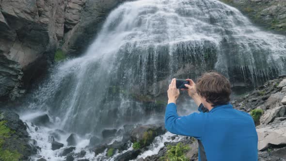 Man Holds Phone and Makes Video Standing Near Waterfall