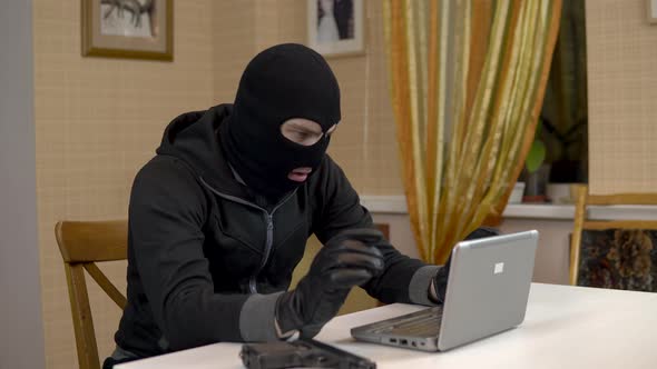 A Robber Is Trying To Hack Into a Laptop. A Masked Thug Is Sitting in a House and Trying To Break