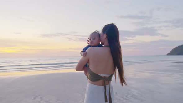 Happy mother with baby boy walks by ocean on the beach in summer