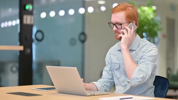 Young Redhead Man with Laptop Talking on Smartphone in Office