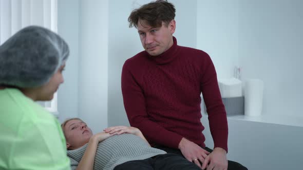 Anxious Worried Caucasian Man Talking with Doctor As Pregnant Woman Lying on Examination Couch in