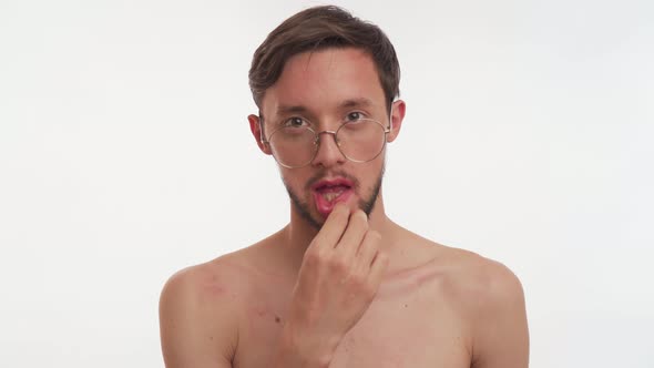 One young Caucasian bearded man stare camera, puts pink lipstick on his lips on white background.