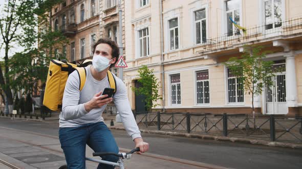 Poartrait of Courier with Yellow Backpack and Protective Mask Rides a Bicycle on the Street Through