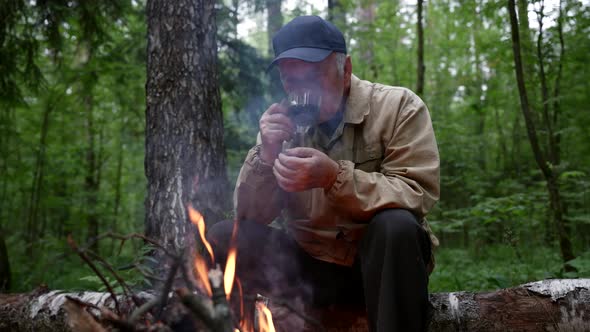 an Elderly Man in a Cap Sits Thoughtfully in the Forest Near a Campfire and Drinks From a Cup