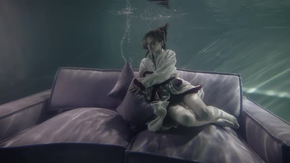 A Model Woman in a Bathrobe Is Swimming and Lying with Pillows on a Gray Sofa That Is Under Water It