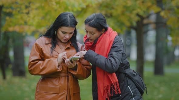 Two Beautiful Hispanic Young Women Stand in Autumn Park Girls Using Smartphone Look at Gadget
