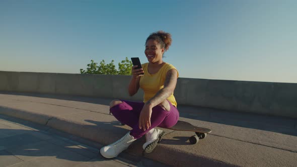 Cheerful Black Female Sitting on Skateboard Online Messaging on Phone
