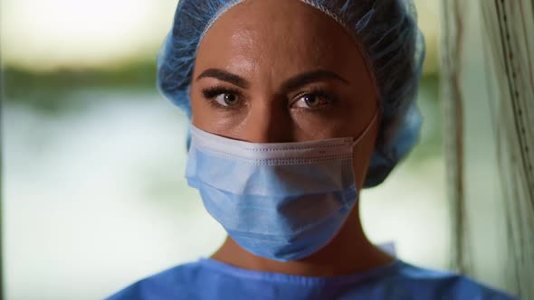 Close View of Young Female Doctor or Nurse Wearing Surgical Face Mask and Cap and Looking at Camera