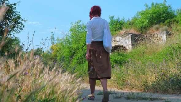 A Red-haired Country Girl Walks Along a Dirt Road or Path in the Countryside