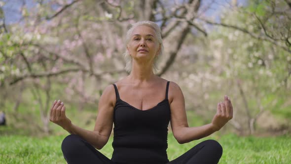 Front View of Confident Fit Senior Sportswoman Sitting in Lotus Pose Meditating in Spring Summer