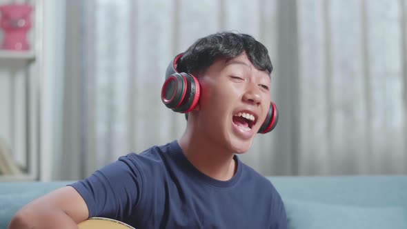 Close Up Of Asian Boy Composer With Notebook On Table Wearing Headphones Singing And Playing Guitar