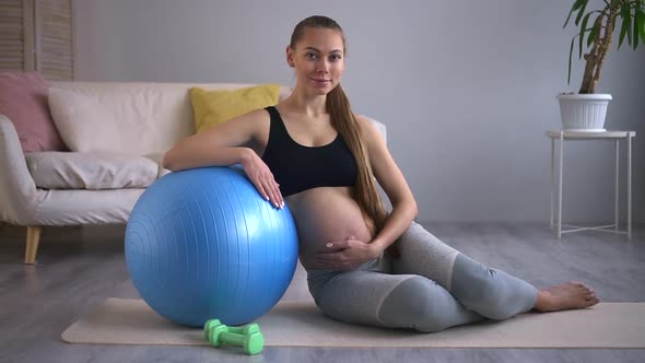 Pregnant Woman Practicing Yoga and Smiling While Sitting on Floor in Home Room Spbd