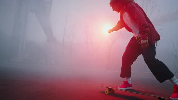 Trendy Teen Guy is Riding a Skateboard in the Evening Holding a Red Burning Torch in One Hand