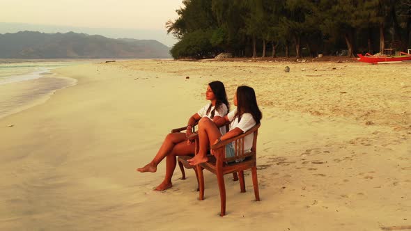 Two girls facing sea, sitting on chairs over white sand of exotic beach on a quiet sunset scenery of