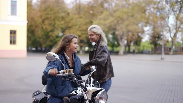 Two Beautiful Young Women Sitting on a Motorcycle at City Street and Smiling