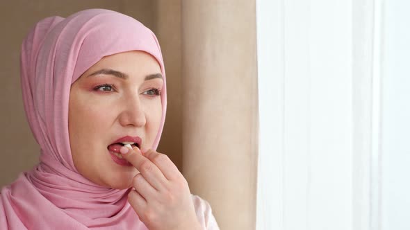 Young Woman in Hijab Drinks White Capsule with Water While Standing By the Window