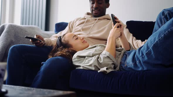 Relaxed Young Couple At Home Sitting On Sofa Watching TV And Checking Social Media On Mobile Phone