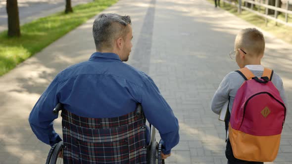 Back View of the Disabled Father Veteran in a Wheelchair and His Son Talk About School and How He