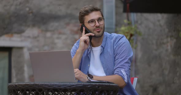 Man Wearing Blue Shirt Working on Laptop Computer and Speaking By Cell Phone While Sitting at Street