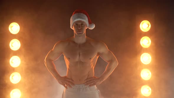 Portrait of Muscular Man Wearing Christmas Santa Hat Showing Finger on Camera on Smoky Background