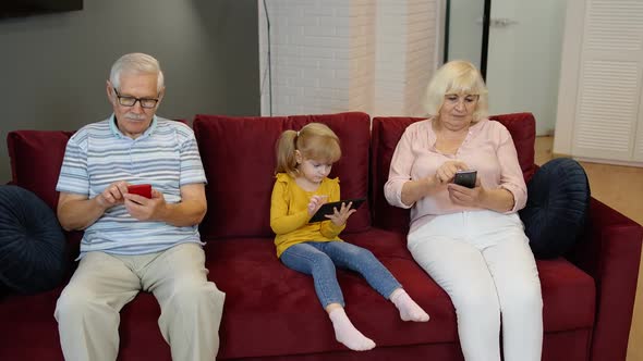 Grandparents and Granddaughter Sit on the Couch at Home and Use Mobile Phones and Digital Tablet