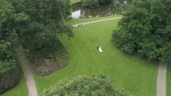 Aerial of Young Wedding Couple Walking on Grass in a Park holding Hands with a Small Pond in the bac