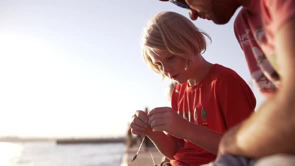 Young Caucasian Blonde Boy in Red Shirt Baiting Fishhook Sitting Near Sea with His Father in