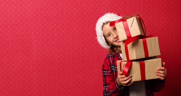 Funny little girl in santa hat with many Christmas gift boxes isolated on red background,