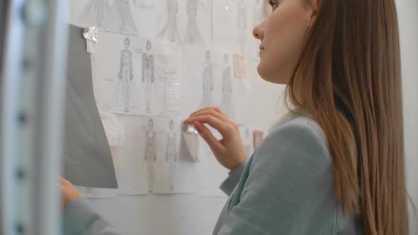 Female Clothes Designer Looking at Drawings of Garments Hanging on Wall