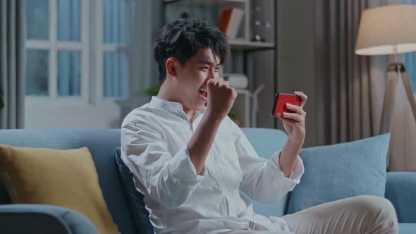 Asian Man Celebrating Winning Game On Smartphone While Sitting On Sofa In The Living Room