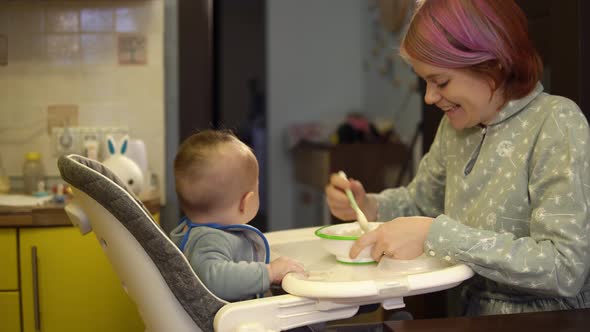 Mother is Feeding Her Son with Porridge By Spoon Sitting in Baby Chair in the Kitchen