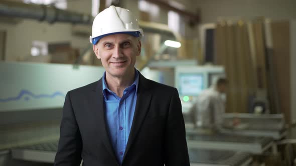 Portrait of Corporation Leader Wearing Helmet Posing on Camera in Front of Workers During Workflow