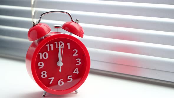 Red alarm clock rang at lunchtime at twelve clock, against the background closed blinds.