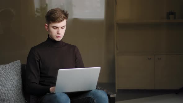 Young Focused Guy Student Sitting on Couch at Home Studying Remotely with Computer During Quarantine