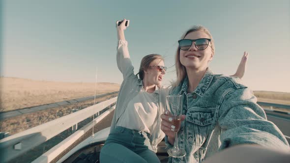 Girl films herself on phone with girlfriend as they ride convertible and drink champagne.