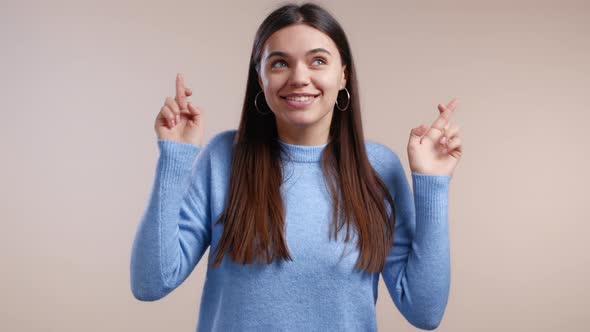 Cute Young Woman in Blue Sweater Praying with Crossed Fingers Over White Background