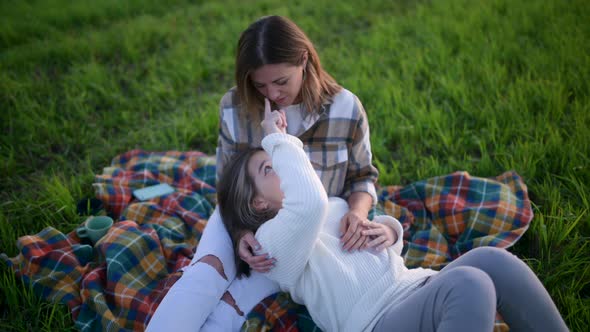 Loving tender mum with a cute daughter sitting on a blanket on a field at sunset