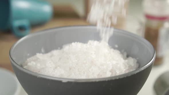 Coarse sea salt is poured into a gray bowl in slow motion on the kitchen table