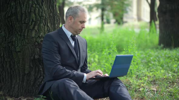 Middle Aged Caucasian Businessman Sitting on Green Meadow Opening Laptop and Typing