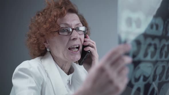 Female Doctor Looking at CT Xray Talking to Colleague on Smart Phone in Hospital Office