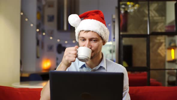 Young man in Christmas santa hat sitting in front of laptop with cup of coffee