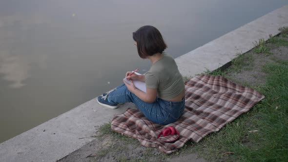 High Angle View of Thoughtful Caucasian Little Woman Writing Diary Sitting on Lake Shore in Summer