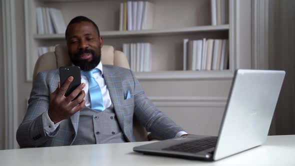 Cheerful African-American Bearded Man in a Gray Suit, White Shirt. The Businessman Is on the Desktop